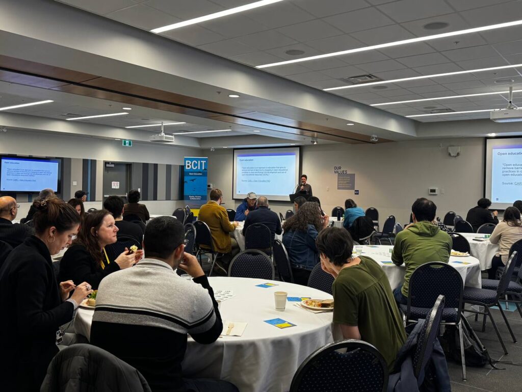A room full of guests seated at tables, with a speaker standing at the front of the room.